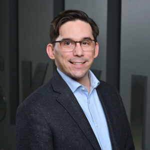 Professional headshot of a man wearing glasses, a blue dress shirt, and a dark textured blazer, smiling in front of a gray wall with partial lettering in the background.